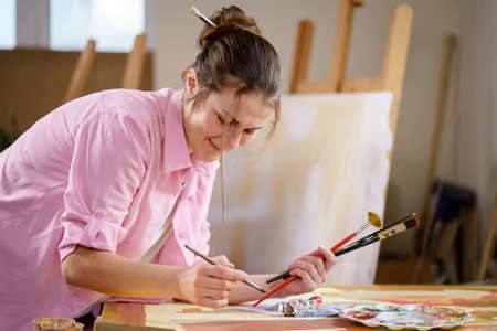 Caucasian woman artist working on a painting in bright daylight studio. Happy artist draws an art project with paints and a brush in the workshop. Hobby. Artist at work. Creative profession.の写真素材