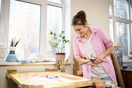 Caucasian woman artist working on a painting in bright daylight studio. Happy artist draws an art project with paints and a brush in the workshop. Hobby. Artist at work. Creative profession.の写真素材