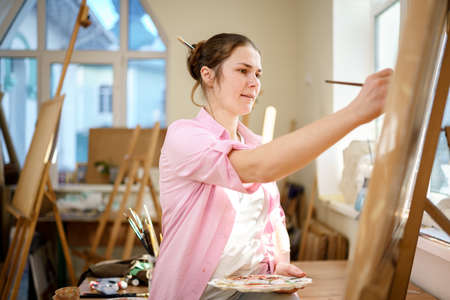 Caucasian woman artist working on a painting in bright daylight studio. Happy artist draws an art project with paints and a brush in the workshop. Hobby. Artist at work. Creative profession.の写真素材