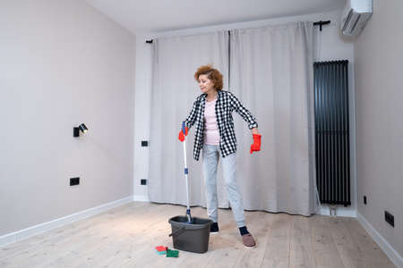 People, housework and housekeeping concept. Happy senior woman in protective gloves cleans floor and dances at home in empty apartment before moving to new home. Excited woman enjoying housekeeping.の写真素材