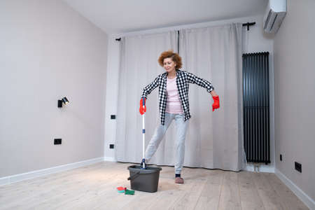 Happy mature woman mops the floor and dances and sings in a new unfurnished apartment before moving to a new house. Housewife enjoying domestic chores, doing home cleanup creatively.の写真素材