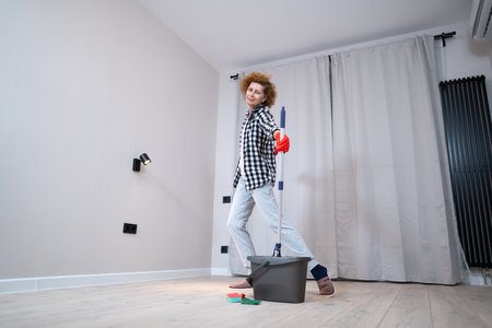 Happy mature woman mops the floor and dances and sings in a new unfurnished apartment before moving to a new house. Housewife enjoying domestic chores, doing home cleanup creatively.の写真素材