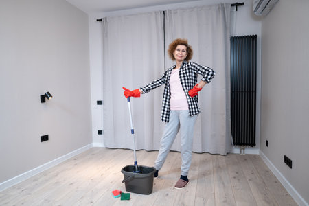 Happy mature woman mops the floor and dances and sings in a new unfurnished apartment before moving to a new house. Housewife enjoying domestic chores, doing home cleanup creatively.の写真素材