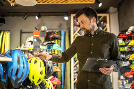 A male bike shop manager makes an inventory of sports helmets in a bike shop. The owner of a sports store with a clipboard in his hands checks the prices of bicycle helmets in the showcase.の写真素材