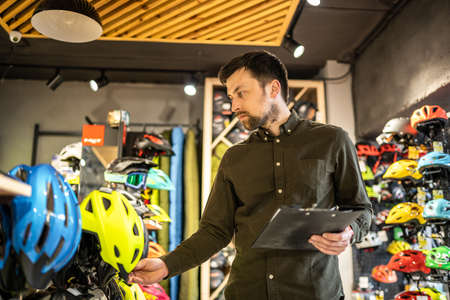 Bike shop manager checks helmet price information on tablet, seller makes an inventory in sports shop. Theme of small business selling bicycles. Seller in hands document checklist in bicycle store.の写真素材