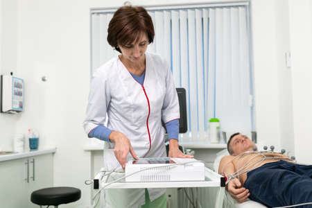 Cardiologist doctor performs electrocardiography procedure on man lying on couch in hospital. ECG test of male at modern clinic. Diagnostic, healthcare, medical service. Hospital patient and EKG test.の写真素材