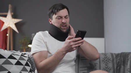 Young man after car accident suffering at home. Male in foam cervical collar reading message on cellphone, feeling pain in neck. Guy wearing neck brace to support neck at home on couch.の写真素材
