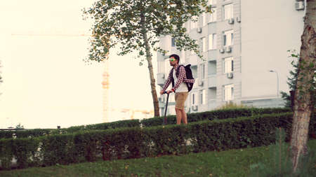 Young man riding on electric scooter on bicycle lane through park. City life. Eco transport. Urban guy ride on e-scooter. Technology, eco friendly mobility concept. Trendy urban transportation.の写真素材