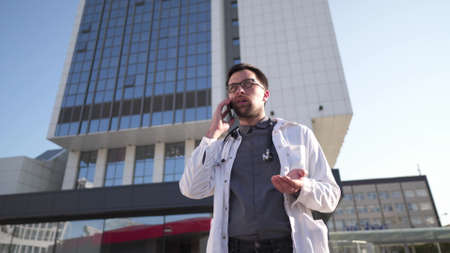 Young Caucasian doctor in white medical coat consults patient on cell phone during break outside hospital. Medical worker talking on mobile phone near clinic. Physician in uniform talk on phone.の写真素材