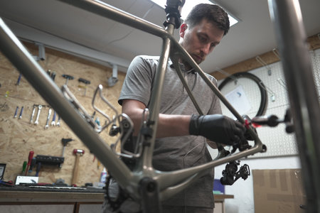 A man is in a workshop, focusing on a bicycle frame. Nearby are automotive tires, bicycle parts, and metal rims. The sound of music adds to the engineering ambiance.の写真素材