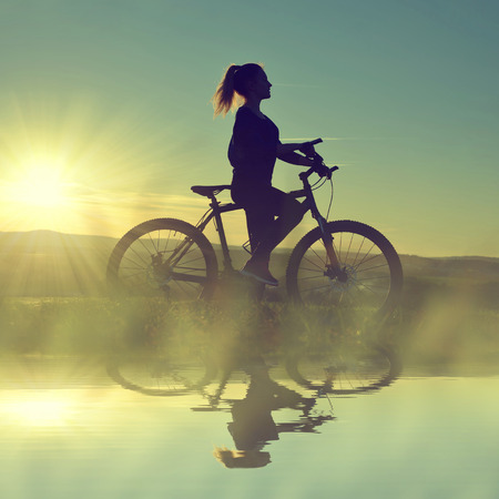 Girl on a bicycle in the sunset reflected on the water surfaceの写真素材