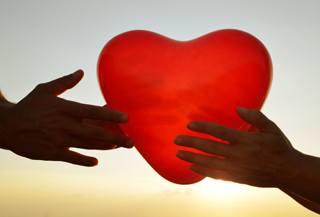 Silhouette of hands holding red balloon in the shape of heart in the background sky with sunlight. Concept of Love.の写真素材