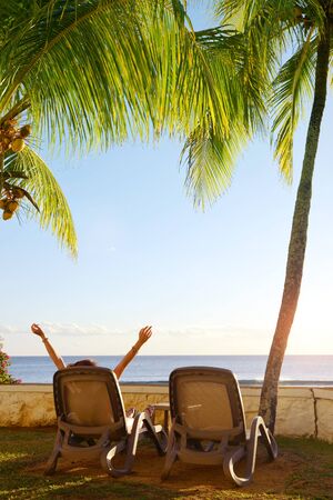 Back view of young woman sunbathing and relaxing under coconut palm tree on Mauritius island tropical beach.の写真素材