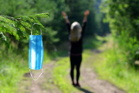 The women on a walk in the woods, in the foreground a medical mask hanging on the branch of spruce tree. Life during covid-19 pandemic.の写真素材