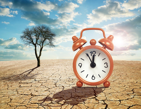 Alarm clock with five minutes before twelve o'clock on arid cracked soil. Concept of climate change or global warming.の写真素材