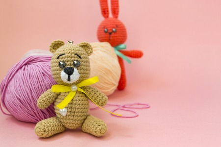 A brown knitted bear sits on a pink background, behind it is a ball of pink and yellow yarn, and an orange knitted hare stands behind the yarn. There is a place for text. The concept of hobby, needlework.の写真素材