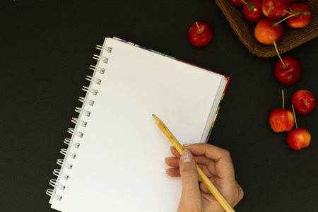 Work space with notebook and red apples on black chalk board background. Female hand holds a pencil and writes plans in the diary. View from above. From above.の写真素材