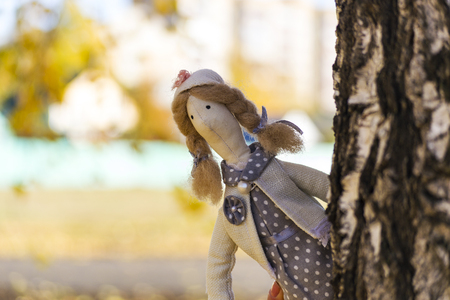 A female Tilda doll in a colorful dress looks out from behind a tree. Registration of holidays. Interior fairy doll handmade. Art and creativity. Blurred background, there is a place for text.の写真素材