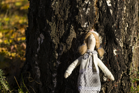 A female Tilda doll in colorful clothes is standing near a birch tree trunk. Registration of holidays. Interior fairy doll handmade. Art and creativity. Blurred background, there is a place for text.の写真素材
