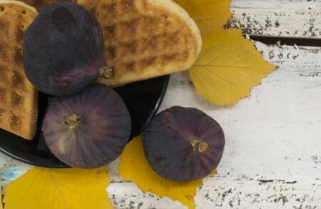 Fig fruits on a plate lying with cookies in the shape of a heart in a plate. Around the plate are the yellow leaves of the trees. The background is white wooden. Autumn concept. There is a place for text.の写真素材
