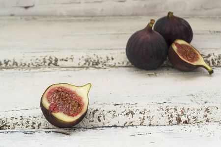 Three fresh figs on a wooden white table. One fig is cut in halves and its flesh is visible. Located...