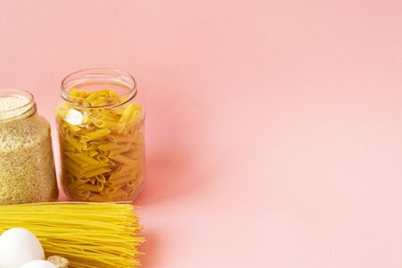 Macaroni, rice are poured into jars, different products and eggs lie on a pink background with place for text. Humanitarian assistance during the coronavirus pandemic. Stock of non-perishable food items.の写真素材