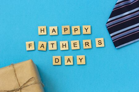 The inscription of wooden letters Happy Father's Day. Flat layout with congratulations. On a blue background are a striped tie, a box in kraft paper.の写真素材