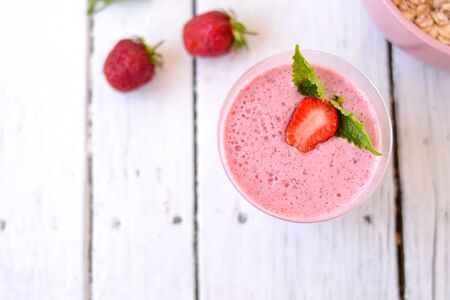 Cocktail with strawberries and milk in a glass decorated with mint and strawberries. Nearby is a pink cup with Hercules flakes, and loose strawberries on a white wooden background. Morning summer breakfast. Healthy breakfast concept.の写真素材
