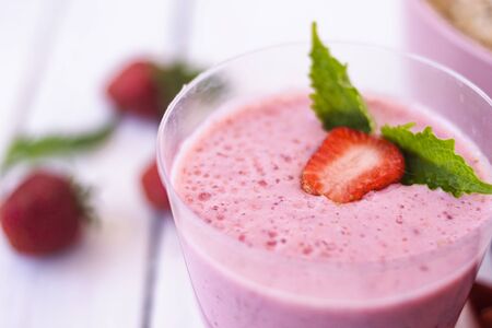 Cocktail with strawberries and milk in a glass decorated with mint and strawberries. Nearby is a pink cup with Hercules flakes, and loose strawberries on a white wooden background. Morning summer breakfast. Healthy breakfast concept.の写真素材