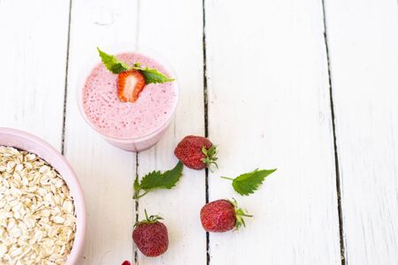 Cocktail with strawberries and milk in a glass decorated with mint and strawberries. Nearby is a pink cup with Hercules flakes, and loose strawberries on a white wooden background. Morning summer breakfast. Healthy breakfast concept.の写真素材