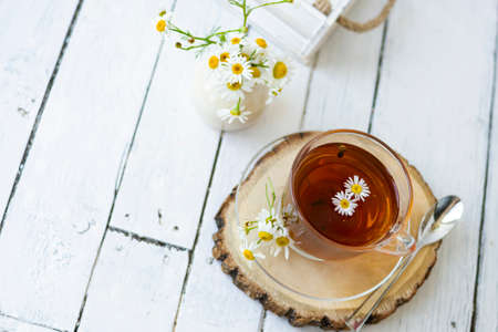 A cup of chamomile and herbal tea with chamomile flowers on white wooden planks. A vase of white flowers stands nearby.の写真素材