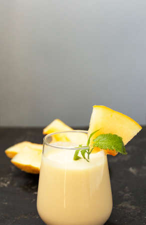 Melon milk shake on a black background. Ripe sliced melon is lying nearby. The smoothie glass is decorated with a sprig of mint and melon crumbs. Summer healthy food concept.の写真素材