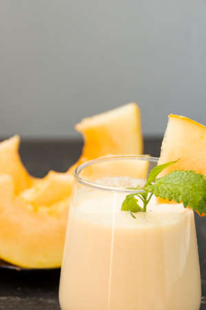 Melon milk shake on a black background. Ripe sliced melon is lying nearby. The smoothie glass is decorated with a sprig of mint and melon crumbs. Summer healthy food concept.の写真素材