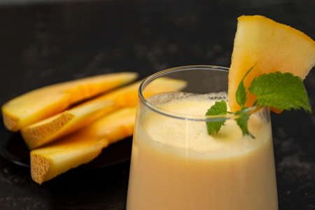 Melon milk shake on a black background. Ripe sliced melon is lying nearby. The smoothie glass is decorated with a sprig of mint and melon crumbs. Summer healthy food concept.の写真素材