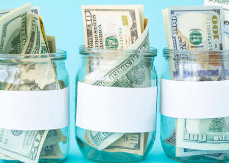 Three Piggy Bank from a Glass Jar for Life. New dollars sticking out of piggy banks on a blue background. The banks have white sheets for inscriptions. Mockup. Copy space. Economy concept.の写真素材