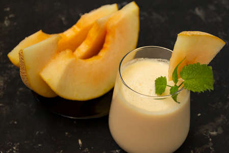 Melon milk shake on a black background. Ripe sliced melon is lying nearby. The smoothie glass is decorated with a sprig of mint and melon crumbs. Summer healthy food concept.の写真素材