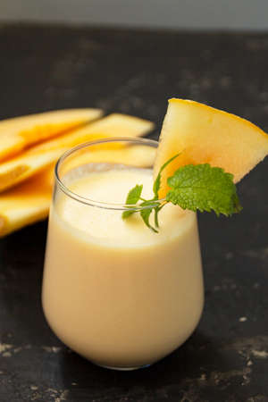 Melon milk shake on a black background. Ripe sliced melon is lying nearby. The smoothie glass is decorated with a sprig of mint and melon crumbs. Summer healthy food concept.