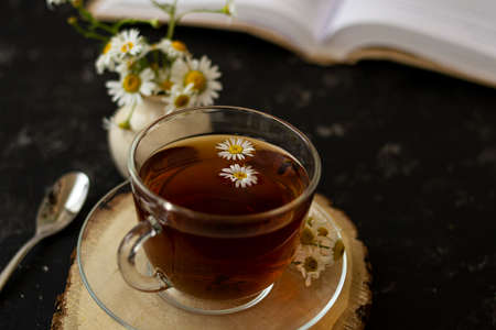 English tea with chamomile stands on a wooden frame and a teaspoon and an open book for reading lies on a black background. Nearby is a white vase with summer chamomile flowers.の写真素材
