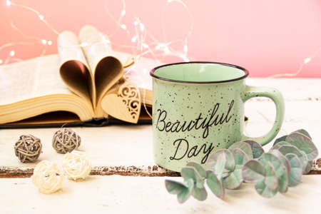 Mint tea mug. On the mug is the inscription of a beautiful day. Nearby lies an open book with heart-shaped pages. Garlands, a eucalyptus branch, decorative balls, and a wooden heart decorate the composition. Pink background.の写真素材