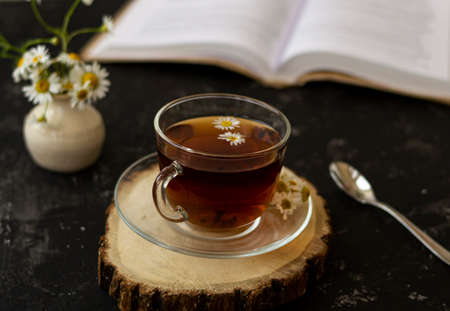 English tea with chamomile stands on a wooden frame and a teaspoon and an open book for reading lies on a black background. Nearby is a white vase with summer chamomile flowers.の写真素材