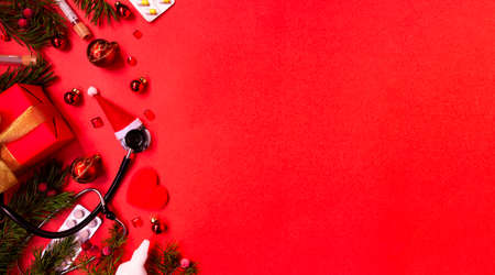 Christmas banner with a Phonendoscope wearing a Santa Claus hat. Nearby lie red balls, a heart, a gift box, branches of a Christmas tree, pills, and medical test tubes. Medical banner concept for New Year. flatly. The colors are red and gold. Copy space.の写真素材