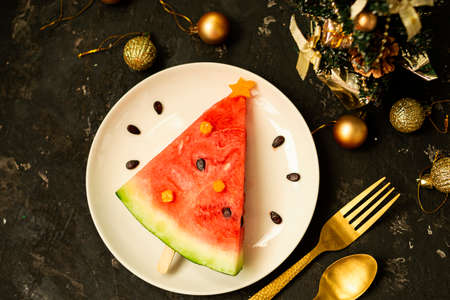 A piece of watermelon is decorated like a Christmas tree on a white plate. Black concrete background with Christmas balls and a fork with a spoon and decor.の写真素材