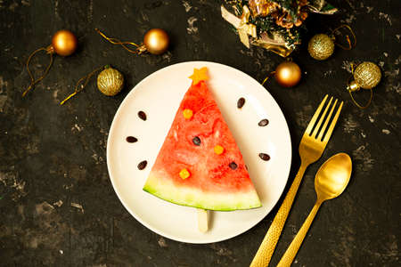 A piece of watermelon is decorated like a Christmas tree on a white plate. Black concrete background with Christmas balls and a fork with a spoon and decor. Breakfast idea for children. New Year, Christmas food with watermelon, top view. celebration.の写真素材