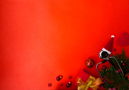 Christmas banner with a Phonendoscope wearing a Santa Claus hat. Nearby lie red balls, a heart, a gift box, branches of a Christmas tree. Medical banner concept for New Year. The colors are red and gold.の写真素材
