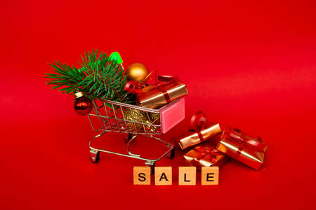 Grocery basket with golden gift boxes and Christmas balls, a sprig of Christmas tree and English letters SALE on a red background banner with copy space. Xmas sale. Christmas shopping online. Winter holiday sales and discounts, seasonal sales, Black Friday, Christmas, discounts and online shopping.の写真素材