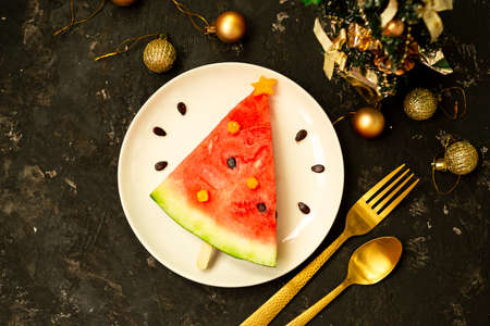 A piece of watermelon is decorated like a Christmas tree on a white plate. Black concrete background with Christmas balls and a fork with a spoon and decor. Breakfast idea for children. New Year, Christmas food with watermelon, top view. celebration.の写真素材