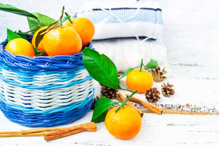 Christmas basket with tangerines, cinnamon sticks and pine cones lie on a wooden white background. Knitted jumpers stack on top of each other and the garland burns beautifully. Hygge concept.の写真素材