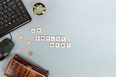 Unemployment concept. A lack of money. Coins fall out of a wallet on a gray background with copy space. The word Unemployment is lined with letters. Computer mouse, keyboard and succulent flower lie nearbyの写真素材