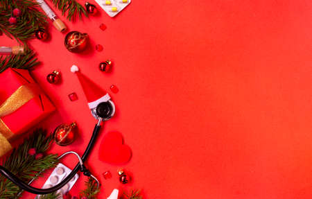 Christmas banner with a Phonendoscope wearing a Santa Claus hat. Nearby lie red balls, a heart, a gift box, branches of a Christmas tree, pills, and medical test tubes. Medical banner concept for New Year. flatly. The colors are red and gold.の写真素材