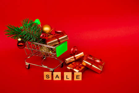 Grocery basket with golden gift boxes and Christmas balls, a sprig of Christmas tree and English letters SALE on a red background banner with copy space. Xmas sale. Christmas shopping online. Winter holiday sales and discounts, seasonal sales, Black Friday, Christmas, discounts and online shopping.の写真素材